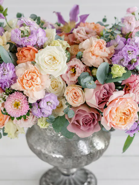 Pastel floral arrangement with roses, carnations, stock, lisianthus, eucalyptus, and seasonal blooms in a silver pedestal vase