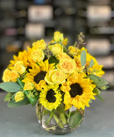 Bright sunflower and yellow rose bouquet in a clear round vase with eucalyptus, handcrafted by Griffin’s Floral Design.