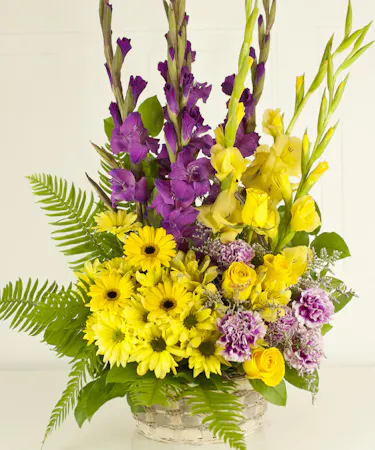 Fresh flowers such as Asiatic lilies, gladioli, carnations and daisy spray chrysanthemums – in shades of yellow and purple – in a woven basket. Approximately 16