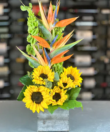Striking floral arrangement with Birds of Paradise, vibrant sunflowers, and lush greenery in a rustic wooden box.